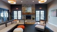 This single-floor space is designed as a sophisticated and inviting lounge area that seamlessly blends comfort with contemporary style. The room's most commanding feature is a floor-to-ceiling natural stone fireplace, flanked by a rustic wooden mantel and framed by a decorative mirror, serving as a bold focal point that anchors the entire space. The deep charcoal-toned walls create a striking contrast against the light-colored stone, lending the room a modern yet warm ambiance. Natural light flows generously through multiple sets of large, divided-light French doors and windows positioned on either side of the fireplace, connecting the interior to the outdoors and brightening the space throughout the day. The seating arrangement is thoughtfully curated, featuring plush sectional and individual sofas in neutral tones accented with orange and grey throw pillows, complemented by bold orange ottoman poufs that add a playful pop of color. Overhead, cylindrical pendant light fixtures with warm orange shades echo the color palette and provide inviting ambient lighting. Recessed ceiling lights further enhance the room's brightness and functionality. The overall atmosphere is energetic yet comfortable, making this lounge well-suited for both social gatherings and relaxed lounging.