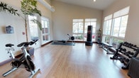 This dedicated fitness studio offers a clean, open layout designed to support a wide variety of workout routines. The space is finished with smooth light wood laminate flooring that provides both durability and a warm aesthetic. Soaring vaulted ceilings amplify the sense of openness and contribute to an airy, energizing atmosphere throughout.

Natural light floods the room through multiple large windows fitted with roller shades, offering views of the surrounding greenery and allowing for excellent ventilation via glass-paneled doors. The bright, neutral wall tones complement the clean and focused ambiance of the space.

The studio is thoughtfully equipped with a professional rowing machine, a freestanding heavy punching bag, a medicine ball rack with multiple weighted balls, a weight bench, and additional cardio equipment positioned throughout the room. Lush potted tropical plants are placed near the entryways, softening the space and adding a refreshing natural element.

Whether used for strength training, cardio, or boxing, this versatile fitness studio provides a well-lit, spacious, and motivating environment suited for comprehensive full-body workouts.
