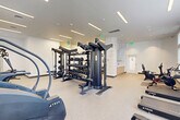 Located at 55 Ridge Lane in Waltham, MA, this fitness center offers a comprehensive, single-floor workout facility designed to support a wide range of exercise routines. The space is bright and welcoming, featuring large windows along multiple walls that flood the interior with natural light and provide views of the surrounding greenery outdoors. The clean, white walls and speckled low-pile flooring contribute to a fresh, modern aesthetic throughout.

The cardio section is well-appointed with a lineup of True-brand treadmills, upright and recumbent stationary bikes, a stair climber, and elliptical machines — each equipped with digital display screens for performance tracking. A dedicated strength training area houses a robust multi-station cable and functional trainer system, a full rack of dumbbells, medicine balls, a power rack with a barbell and weight plates, and an adjustable bench, providing ample options for both beginner and advanced lifters. Additional strength machines, including a leg press unit, round out the equipment selection.

Recessed and linear LED lighting supplements the natural light throughout the space, ensuring consistent visibility. The facility is finished with a dedicated water station, safety signage, and a direct exterior entry door for convenient access. The overall layout is thoughtfully organized to maximize the usable workout area while maintaining clear, open circulation paths between equipment zones.