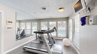This single-floor fitness and wellness facility offers a well-organized layout designed to support an active lifestyle. The main exercise room is bright and inviting, with multiple windows fitted with slatted blinds that allow generous natural light while offering views of the surrounding outdoor area. Large wall mirrors visually expand the space, and a textured ceiling adds a classic finish overhead. The room accommodates cardio equipment, including treadmills and a stationary bike, alongside a stability ball, making it a versatile workout environment. A wall-mounted television provides entertainment during exercise sessions.

Adjacent to the fitness area is a traditional sauna, enclosed by a wood-framed frosted-glass door and finished with warm, rich wood-paneled walls and tiered benching — ideal for relaxation and recovery.

The two bathrooms are finished with neutral square floor tiles and half-wall ceramic tile wainscoting, giving them a clean, functional appearance. Each features standard fixtures, vanity mirrors with overhead lighting, and enclosed shower areas with glass doors. The tiled flooring and walls ensure easy maintenance and a hygienic environment. Together, these spaces form a cohesive, amenity-focused facility well-suited for community or residential use.