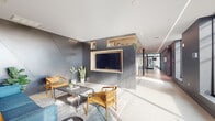 This single-floor amenity space showcases a cohesive contemporary design throughout its key areas. The lounge area serves as a welcoming centerpiece, featuring dark paneled walls with angular geometric detailing, warm wood built-in shelving, and a wall-mounted television. Comfortable seating arrangements, including leather-accented chairs and a vibrant blue sofa, create inviting spaces for both relaxation and casual work. Integrated workstations with sculptural wood partitions offer semi-private areas ideal for focused tasks. Large floor-to-ceiling windows line the corridors and fitness areas, flooding the space with natural light and framing views of the surrounding streetscape and distant skyline. The elevator lobby is defined by sleek, dark metal wall paneling and linear LED ceiling lighting that runs the length of the hallway, lending a polished, modern aesthetic. The expansive fitness center is well-equipped with a full range of strength machines, cardio equipment including treadmills and ellipticals, free weights, and stability balls, complemented by floor-to-ceiling mirrors and a bold pop-art mural as a focal accent. A dedicated stretching and flexibility area provides additional space for varied workouts. Together, these areas present a refined, functional amenity floor with a consistent modern design language.
