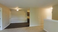 This single-floor residence offers a clean, functional layout designed for comfortable everyday living. The main living area features plush carpeting that adds warmth underfoot, while an adjacent section transitions to rich, dark hardwood-style flooring, creating a clear visual distinction between living and dining or transitional spaces. Neutral, light-toned walls throughout lend the interior a bright and airy feel, complementing the natural and artificial lighting. A ceiling fan with an integrated light fixture is centrally positioned in the living area, providing both comfort and illumination. The layout flows openly toward the kitchen, which is partially visible and showcases dark espresso-toned cabinetry paired with light countertops, hinting at a well-designed culinary space. A low partial wall or bar-height counter at the edge of the living area subtly defines the boundary between spaces without closing off the open feel. White baseboards and clean wall finishes add a crisp, polished detail throughout. The overall ambiance is neutral and move-in ready, offering a versatile canvas well-suited for a variety of interior design styles.