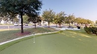 Located at 5533 Houston Rd in Macon, Georgia, this outdoor putting green offers a well-designed recreational space for golf enthusiasts and casual players alike. The green features multiple putting holes with flag markers distributed across a gently contoured synthetic turf surface, providing varied challenge and engagement for users of all skill levels. Surrounding the green are carefully maintained sand trap areas, adding an authentic golf course feel to the amenity.

The space is beautifully framed by mature trees, ornamental sago palms, and manicured shrubbery, creating a lush, park-like atmosphere. The landscaping provides both shade and visual appeal, enhancing the overall experience of the green. A gravel or crushed-stone border defines the perimeter of the putting surface, neatly separating it from the surrounding greenery.

Adjacent to the putting green, a picnic area with tables offers a convenient spot for relaxation and socializing. Views from the green reveal a nearby pool area complete with a covered pavilion and umbrella-shaded seating, suggesting this putting green is one of several outdoor amenities within the community. The broader grounds are lined with well-spaced trees and open lawns, contributing to a peaceful, resort-like environment under the open Georgia sky.