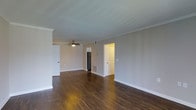 This single-floor space presents a clean, open layout with a cohesive and practical design. The main living area is finished with rich, dark hardwood flooring that extends throughout, providing a warm contrast against the neutral gray walls. Crown molding runs along the ceiling perimeter, adding a subtle architectural detail that elevates the overall finish of the space. A ceiling fan is centrally mounted, contributing both functionality and a modest decorative element.

The open sightlines from the main area allow views into adjacent spaces, including what appears to be a kitchen equipped with a stainless steel refrigerator, as well as access to additional rooms through white paneled doors. The white baseboards and trim throughout provide clean definition between the flooring and walls, reinforcing a tidy, well-maintained aesthetic.

The layout is straightforward and efficient, with a visible floor vent indicating a central HVAC system. The overall ambiance is neutral and move-in ready, with a palette and finish selection that suits a variety of furnishing styles. Natural light appears to supplement the interior lighting, contributing to a bright and functional environment.