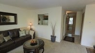 This single-story space features a comfortable living room with plush wall-to-wall carpeting and neutral cream-toned walls that create a warm, inviting atmosphere. The living area is furnished with a dark leather sofa accented with decorative pillows and anchored by a round glass-topped coffee table with a dark wood base. A notable architectural detail is the pass-through window cut into the dividing wall, which offers a direct view into the kitchen, where a refrigerator and cabinetry are visible, enhancing the open feel between the two spaces. Decorative elements include a tall branching arrangement in a stylized planter and a table lamp that adds warm ambient lighting. A hallway extends from the living area toward additional rooms, where natural light filters in through a window at the far end, brightening the passage. A dark area rug near the hallway entry marks a transition from the carpeted living space. The overall ambiance is understated and functional, with a clean, neutral palette throughout.