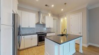Located at 1414 Broadview Ave in Columbus, Ohio, this single-story residence offers a thoughtfully designed interior with clean, contemporary finishes. The kitchen stands as a highlight of the home, showcasing crisp white shaker-style cabinetry that extends to the ceiling, complemented by dark quartz countertops that provide a striking contrast. A central island with an undermount sink and matching dark countertop offers additional prep space and casual seating, illuminated by elegant glass pendant lights that add a refined decorative touch. The stainless steel range is flanked by a sleek wall-mounted hood, while a light blue subway tile backsplash introduces a subtle pop of color that ties the space together. Recessed lighting throughout ensures the kitchen is well-lit and functional. Crown molding adds a classic architectural detail to the ceilings, enhancing the overall sense of quality and craftsmanship. Light hardwood flooring runs through the space, warming the interior and providing a natural contrast to the white cabinetry. A doorway off the kitchen provides a glimpse into an adjacent room, suggesting an open and connected floor plan. The overall ambiance is modern, clean, and inviting.