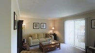 This single-floor residence offers a thoughtfully arranged living space defined by a warm and inviting atmosphere. The living room serves as the heart of the home, finished with wall-to-wall carpeting and painted in soft, neutral tones that create a calm and welcoming ambiance. A large sliding glass door fitted with vertical blinds allows an abundance of natural light to fill the room while providing access to the outdoors. The space is well-proportioned, comfortably accommodating a plush sofa accented with decorative pillows and a classic wooden coffee table featuring a glass top and lower storage shelf. Subtle accent lighting from floor and table lamps adds warmth to the room during evening hours. Framed artwork adorns the walls, contributing to the refined yet understated aesthetic of the interior. Crown molding along the ceiling line adds an elegant architectural detail, enhancing the overall finish of the space. The layout is practical and functional, with smooth transitions between areas that make the floor plan feel cohesive and easy to navigate. Overall, this residence presents a clean, move-in-ready environment with classic design elements that appeal to a wide range of tastes.