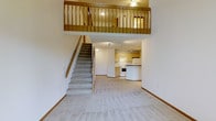 This two-story residence is designed with an open and airy layout that immediately draws the eye upward to the dramatic double-height ceiling in the main living area. A carpeted staircase with a warm wood handrail leads to the upper level, where a loft-style landing overlooks the main floor below, framed by a classic wooden baluster railing. The open-concept main level seamlessly connects the living area to the kitchen, which features light wood cabinetry, a white appliance suite including a range and refrigerator, and a curved peninsula that provides additional counter space and serves as a casual dining or prep area. Neutral-toned carpeting throughout the main floor lends a consistent, comfortable feel to the space, while the clean white walls offer a bright, move-in-ready canvas. The upper loft level, illuminated by a ceiling fan light fixture, provides flexible space suitable for a bedroom, home office, or additional living area. Beyond the interior, the property enjoys serene outdoor views of lush greenery, mature evergreen and deciduous trees, and natural open land with seasonal wildflowers, providing a peaceful natural backdrop visible from the exterior of the building.