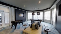 This single-floor entertainment and lounge space is thoughtfully designed to serve as a premier social hub. The room features a distinctive curved wall of windows that floods the interior with natural light, framed by elegant draping curtains. The flooring consists of a geometric, multi-toned blue and gray carpet that adds visual depth and texture throughout the space.

A wall-mounted flat-screen television is set against a bold, dark charcoal accent wall, creating a striking focal point that anchors the room. A comfortable, contemporary sofa with decorative throw pillows is centrally positioned, complemented by sleek, minimalist side tables in a dark wood finish. Two woven, globe-shaped pendant lights hang above the pool table area, adding warmth and an artistic contrast to the otherwise modern aesthetic.

For recreation, the room includes a full-size pool table positioned near the windowed alcove and a foosball table on the opposite side, making the space well-equipped for entertainment. Recessed ceiling lighting provides supplemental illumination. The overall ambiance strikes a balance between sophisticated design and casual, recreational functionality.