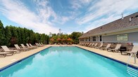 This single-story community amenity space is designed to support an active lifestyle, offering both indoor fitness facilities and an inviting outdoor pool area. The outdoor pool is generously sized, featuring crystal-clear turquoise water surrounded by a wide concrete deck lined with lounge chairs. Vibrant red market umbrellas provide shaded seating areas, while mature trees form a lush green backdrop, creating a resort-like atmosphere under open skies.

The indoor fitness center is thoughtfully laid out across two connected areas. The main exercise room features vaulted ceilings that lend a sense of openness and airiness, with large windows along the walls flooding the space with abundant natural light and views of the surrounding greenery. Floor-to-ceiling mirrors along one wall visually expand the room and allow exercisers to monitor their form. The space is equipped with multiple treadmills, stationary bikes, elliptical trainers, a cable weight machine, a dumbbell rack, and an adjustable workout bench, providing a comprehensive range of cardio and strength training options. A wall-mounted television and a clock complete the practical amenities. The entry area features a welcoming foyer with a reception-style counter, offering a clean and organized transition into the workout space. Neutral warm tones throughout the interior create a calm, focused environment.