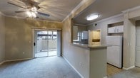 This single-floor space presents an open and functional layout that integrates the living area and kitchen seamlessly. The living room is finished with plush carpeting and features a ceiling fan with lighting, crown molding, and a textured ceiling that adds subtle architectural character. Large sliding glass doors dominate one wall, allowing natural light to fill the room while providing direct access to an outdoor balcony with views of the surrounding neighborhood.

Adjacent to the living area, a peninsula-style kitchen counter with a granite-look surface serves as a natural divider between the two spaces. The kitchen is equipped with white cabinetry offering ample storage, a built-in range, and a refrigerator. A dedicated overhead light fixture illuminates the kitchen workspace, while crown molding continues throughout, maintaining visual consistency across the space.

The warm, neutral wall tones throughout create a cohesive and inviting ambiance. The combination of natural light from the sliding doors, practical finishes, and an open floor plan makes this space feel welcoming and well-connected.