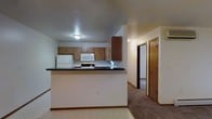 This single-floor space is designed with a functional, open layout that connects the kitchen to the rest of the unit. The kitchen is equipped with medium-toned wood cabinetry offering ample storage and is outfitted with white appliances, including a refrigerator, electric range, and built-in microwave. A peninsula with a dark laminate countertop serves as a natural divider between the kitchen and the adjoining areas, providing additional workspace and a casual dining or serving surface. The kitchen flooring features light-toned vinyl, creating a practical surface for the cooking area. Transitioning from the kitchen, the space opens into a carpeted hallway that leads to additional rooms, each accessible through solid wood doors with matching trim. The warm wood tones of the cabinetry and doors lend a cohesive, traditional aesthetic throughout. Flush-mount ceiling lighting provides consistent illumination across the space. The overall layout is straightforward and efficient, making good use of the available footprint while maintaining clear sightlines between the kitchen and the rest of the living areas.
