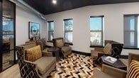 This single-floor amenity space blends industrial and contemporary design elements to create a sophisticated, multi-functional environment. The entrance foyer sets a refined tone with light wood-look tile flooring, bold abstract artwork, and sleek glass-paneled doors leading into the conference room — clearly marked and thoughtfully separated for privacy and focus.

The conference room is a standout feature, anchored by a solid wood dining table surrounded by a mix of chevron-patterned upholstered chairs and leather seating. An ornate gold globe chandelier hangs overhead, complementing the dark ceiling and exposed silver ductwork that add industrial character. Decorative gear wall art and patterned drapery frame large windows that bring in natural light, while a wall-mounted flat-screen TV enhances the room's functionality.

Adjacent to the conference room, a comfortable lounge area offers plush seating arrangements on a bold chevron cowhide rug, surrounded by multiple windows with views of the outdoors. The dark ceiling and exposed ductwork continue here, lending visual cohesion across the space.

A well-appointed coffee bar station features striking black and gold geometric tile backsplash, dark marble countertops, white cabinetry, and a Starbucks-branded coffee machine — providing a premium refreshment experience. Light wood-look flooring flows through the lounge and service areas, tying the overall design together seamlessly.