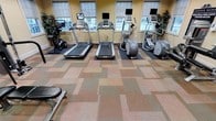 This fitness room offers a clean, functional environment designed for cardiovascular training. The space is finished with multi-toned modular carpet tiles in earthy shades of beige, rose, and sage, lending a warm, organized aesthetic to the floor. Multiple large windows line the walls, fitted with roller shades that allow ample natural light to fill the room while maintaining privacy. The warm yellow-toned walls contribute to an inviting atmosphere.

The equipment lineup includes two treadmills, two elliptical cross-trainers, and a stationary cycle, each equipped with individual monitor screens for entertainment or workout tracking. The machines are positioned on protective rubber mats, keeping the flooring in good condition. Potted plants are placed near the windows, softening the space and adding a touch of greenery.

The layout is thoughtfully arranged to provide sufficient clearance between each machine, ensuring comfortable use during peak times. The overall ambiance is bright, approachable, and practical, making this fitness room a convenient amenity for residents or guests seeking a dedicated space for daily cardio exercise.