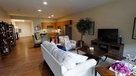 This single-floor residence offers an open-concept design that integrates the living, dining, and kitchen areas into one cohesive and functional space. The living area is anchored by a plush white sofa and complemented by a patterned accent chair, creating a comfortable and inviting atmosphere. Rich hardwood flooring flows throughout, adding warmth and continuity to the space. Recessed lighting provides consistent, well-distributed illumination across the open floor plan. A large decorative tree adds a touch of greenery, enhancing the natural ambiance of the room. The entertainment area features a flat-screen television atop a dark wood media console. The kitchen is outfitted with warm honey-toned cabinetry, stainless steel appliances including a full-size refrigerator, and a tasteful tile backsplash that adds visual interest. Pendant lighting above the kitchen area adds a refined touch. Adjacent to the kitchen, the dining area accommodates a table and chairs, making it ideal for both casual meals and entertaining. The overall ambiance of the space is warm, modern, and thoughtfully curated, offering a balanced blend of comfort and style.