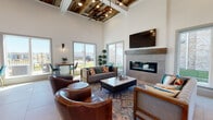 This single-story clubhouse blends rustic and contemporary design elements to create a welcoming, multi-functional gathering space. The main lounge area commands attention with dramatically tall ceilings adorned by a coffered section finished in reclaimed wood planking, complemented by an exposed ceiling fan and pendant lighting that cast a warm, inviting glow. A sleek, linear gas fireplace with a concrete tile surround and a solid wood mantel serves as a striking focal point, topped by a wall-mounted television. Generous seating, including tufted leather armchairs and a fabric sofa accented with colorful throw pillows, is arranged over a richly patterned area rug, encouraging relaxed conversation.

Large sliding glass doors and oversized windows line the perimeter, flooding the interior with abundant natural light while framing views of the surrounding outdoor greenery. Light-toned tile flooring runs throughout, lending a clean, cohesive look. A bistro-style high-top table with contemporary chairs provides an intimate seating nook bathed in sunlight. The open floor plan also accommodates a billiards table and a casual dining area, offering diverse entertainment options. A dedicated beverage station features granite countertops, subway tile backsplash, and a self-serve coffee and water machine, adding a hospitality-forward amenity to the space.