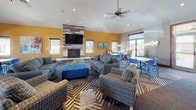 This single-floor community clubhouse is thoughtfully designed to serve as a welcoming hub for socializing and relaxation. The main lounge area immediately captures attention with its vaulted ceilings, bold yellow accent walls, and a striking floor-to-ceiling fireplace clad in rustic reclaimed wood-style tile, topped with a large wall-mounted flat-screen television. A ceiling fan overhead and an abundance of natural light from multiple windows contribute to the bright, airy atmosphere throughout the space.

The lounge is furnished with generous seating arrangements, including gray sofas, patterned accent chairs, and a vivid blue tufted ottoman, all anchored by a bold geometric-patterned area rug. Multiple round dining tables with blue chairs are positioned nearby, offering flexible space for casual meals or group activities.

Adjacent to the main lounge, a fully equipped kitchen provides rich warm-toned cabinetry, granite countertops with a subtle tile backsplash, black appliances including a double wall oven and a side-by-side refrigerator, and a large island with a granite surface and yellow bar stools. Large-format tile flooring adds a clean, polished finish to the kitchen area. Together, these spaces create a functional and stylish environment suited for community events and everyday gathering.