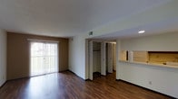This single-floor unit is designed with an open, functional layout that maximizes the available living space. The main living area is finished with rich, dark hardwood-style flooring that extends throughout, providing a warm and cohesive aesthetic. A large sliding glass door fitted with vertical blinds dominates one wall, allowing natural light to fill the space while offering direct outdoor access and views of the surrounding greenery.

A key organizational feature of the unit is the set of bifold closet doors positioned along the interior wall, revealing a spacious built-in storage area complete with a hanging rod and shelving — offering practical, well-integrated storage solutions. Adjacent to this, a pass-through counter connects the living area to the kitchen, promoting an open flow between the two spaces while maintaining a degree of separation. The kitchen area, visible through the pass-through, includes overhead cabinetry.

The ceilings feature a textured finish and incorporate recessed HVAC vents, ensuring comfortable climate control throughout. Neutral-toned walls complement the flooring and contribute to a clean, move-in-ready atmosphere suited for a variety of furnishing styles.