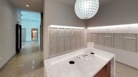 The mail room is a thoughtfully designed communal space that balances functionality with refined aesthetics. The room's most prominent feature is a large, marble-topped central island with built-in drop slots, providing a practical surface for sorting and managing correspondence. Warm wood paneling on the island's base adds a contrasting material detail that complements the room's neutral palette.

Lining the walls are extensive rows of individual tenant mailboxes finished in a brushed silver-grey tone, accommodating a large number of residents. A dedicated section of oversized package lockers, also finished in matching tones, provides secure storage for parcel deliveries. Continuous LED strip lighting installed along a perimeter ledge near the ceiling casts an even, ambient glow throughout the space, enhancing visibility and contributing to the room's clean, modern feel.

Overhead, a distinctive globe pendant light serves as a focal point, adding an elegant design touch to an otherwise utilitarian space. The flooring is a polished terrazzo, reinforcing the contemporary and durable character of the room. An adjacent corridor, visible through the space, connects the mail room to the broader building, ensuring convenient access for residents.
