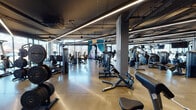 This contemporary fitness facility delivers a premium workout experience within a thoughtfully designed, open-plan layout. The space features an industrial-inspired aesthetic, with exposed ceilings showcasing structural beams, ductwork, and sleek linear LED lighting that illuminates the polished concrete floors throughout.

The facility is organized into distinct training zones. A dedicated strength area houses a comprehensive selection of selectorized machines, free weights, barbells, squat racks, and adjustable benches, complemented by vibrant geometric murals that energize the environment. A separate functional training zone features a cable rig system, weighted sleds, medicine balls, and floor mats, anchored by floor-to-ceiling windows that frame sweeping outdoor views and flood the space with natural light.

The cardio section is lined with a row of treadmills and additional cardiovascular equipment, positioned along the windowed perimeter to maximize natural daylight and outdoor sightlines. Concrete support columns define the space architecturally while maintaining an open, airy feel.

A striking large-scale mural depicting a mountain climber serves as a focal point near the functional training area, reinforcing an active and adventurous atmosphere. The combination of abundant natural light, high ceilings, modern equipment, and bold artistic accents creates a motivating and professional fitness environment.
