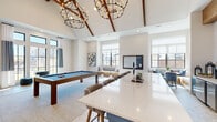 Located at 15901 Championship Pkwy in Fort Worth, TX, this game room is a thoughtfully designed entertainment space that combines modern aesthetics with inviting recreational amenities. The room is defined by its dramatic vaulted ceiling with exposed wood beam trusses, creating an airy, open atmosphere that feels expansive and welcoming. Two statement chandelier fixtures with candle-style lighting anchor the space and complement the warm, contemporary design.

At the heart of the room sits a full-size pool table with a slate-blue felt surface, surrounded by ample open floor space for play. A shuffleboard table is positioned along a striking natural wood accent wall, which also frames a mounted flat-screen television, making it a focal point of the room. Adjacent to the shuffleboard area is a well-appointed wet bar with navy blue cabinetry, a white tile backsplash, and seating along a quartz-topped island that extends into the space.

Large black-framed windows line the walls, flooding the room with natural light and offering views of the surrounding landscape. Built-in window bench seating provides relaxed lounge areas, and additional casual seating with side tables is spread throughout. A dedicated board game display and chalkboard wall add further interactive elements, making this game room a comprehensive and vibrant hub for social entertainment.