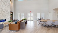 This well-appointed single-story community amenity space delivers a thoughtfully designed environment across several distinct functional areas. The centerpiece of the space is a grand lounge featuring dramatically soaring double-height ceilings, a floor-to-ceiling stacked stone fireplace flanked by built-in shelving, and elegant herringbone-patterned tile flooring. Abundant natural light pours in through large windows and clerestory windows above, creating a bright and inviting atmosphere. Comfortable seating arrangements with leather and upholstered furniture invite relaxation around the fireplace.

Adjacent to the lounge, a fully equipped kitchen serves as a social hub, complete with dark espresso cabinetry, granite countertops, a subway tile backsplash, stainless steel appliances, and a large island with bar seating beneath pendant lighting. A dedicated leasing or administrative office area connects seamlessly via the herringbone-floored corridor.

The business center offers a quiet, carpeted workspace lined with tall windows and multiple work tables ideal for productivity. The fitness center rounds out the amenities with rubber flooring, a ceiling fan, and a comprehensive selection of cardio and strength-training equipment, all bathed in natural light from surrounding windows.