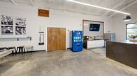 Located at 1 Catawba Cir in Columbia, SC, this single-floor amenity space is a thoughtfully designed, open-plan gathering hub that blends industrial architecture with modern comfort. Soaring vaulted ceilings with exposed white-painted trusses and ductwork create an airy, expansive atmosphere throughout, while polished concrete floors tie the entire space together with a sleek, contemporary aesthetic.

At the heart of the space stands a striking double-sided fireplace clad in warm wood tile, serving as both a functional focal point and a dramatic architectural statement. Surrounding it, the open layout is divided into distinct zones: a lounge area with a large sectional sofa, colorful accent pillows, a mounted flat-screen TV, and a tiled feature wall; a dining and meeting area with a natural wood table and modern chairs; and a recreation zone featuring a pool table.

Floor-to-ceiling black-framed windows line the exterior walls, flooding the interior with natural light and offering views of the lush, palm-tree-lined surroundings. A kitchenette with white cabinetry, a Pepsi vending machine, and a mounted TV provides convenience, while a bank of parcel lockers near the entrance adds a practical amenity. The warm wood-paneled ceiling sections and contemporary lighting throughout complete this inviting, multi-use community space.