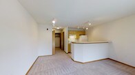 This single-floor space is designed with an open-concept layout that seamlessly connects the kitchen and living area, creating a cohesive and functional environment. The kitchen is equipped with light wood cabinetry, a white refrigerator, and a white range, all illuminated by a track lighting system mounted on the ceiling. A curved breakfast bar with a white finish and wood-toned trim serves as a natural dividing point between the kitchen and the living area, offering a convenient surface for dining or casual use. The living area features neutral-toned wall-to-wall carpeting that adds warmth and comfort underfoot, complemented by clean white walls and baseboards with matching wood-toned trim throughout. Track lighting extends into the living space, ensuring consistent illumination across the open floor plan. A hallway leads to additional areas of the unit, including a walk-in closet with visible shelving, providing practical storage solutions. The overall ambiance is bright, clean, and understated, with a neutral color palette that lends itself well to a variety of interior styles. The layout makes efficient use of space while maintaining an open and airy feel throughout.