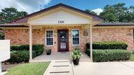 This single-story building presents a classic and professional exterior characterized by warm brick masonry and crisp white trim. The structure features a covered front porch supported by a gabled roofline, creating an inviting and sheltered entryway. A deep burgundy glass-paneled door serves as the focal point of the facade, flanked symmetrically by two white-framed windows dressed with blinds, lending the building a balanced and orderly appearance.

The covered porch extends over a concrete walkway, providing a functional transition from the exterior grounds to the entrance. Decorative potted plants, including a striking flowering arrangement and a tall ornamental grass, flank the doorway and add a touch of color and life to the entry. A professional signage plaque is mounted near the entrance, indicating the building's commercial or professional use.

The surrounding grounds are well-maintained, with neatly trimmed hedgerows lining the foundation and lush green lawn areas framing the approach. Mature trees visible in the background contribute to a serene, established setting. The overall aesthetic is clean, approachable, and professional, suitable for a business or office environment.