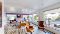 Located at 6649 Ridge Avenue in Philadelphia's Roxborough neighborhood, this compact yet efficiently designed single-floor space offers a thoughtfully curated layout with a combined living and dining room and a fully equipped kitchen. The open main area is defined by an abundance of natural light streaming through large, floor-to-ceiling windows that line multiple walls, offering views of the surrounding streetscape and landscaping. Crisp white walls contrast with a bold accent wall painted in a warm lavender-purple hue, which anchors the space and adds a distinct personality. Rustic wood-look flooring runs throughout, lending warmth and continuity between areas. The living zone features comfortable seating arrangements, while a dedicated workspace area with desks and task chairs makes the space highly functional. A sleek wall-mounted fireplace and decorative circular wooden wall art add stylish focal points. The kitchen is finished with white shaker-style cabinetry, dark granite countertops, a classic white subway tile backsplash, stainless steel appliances, and a deep undermount sink — delivering a clean, modern aesthetic. Recessed lighting complements the natural light, ensuring the space remains bright and welcoming throughout the day.
