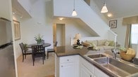 This two-floor space offers a thoughtfully designed open-concept layout that seamlessly connects the kitchen, dining, and living areas into one cohesive and inviting environment. The kitchen is outfitted with crisp white cabinetry, dark granite-style countertops, a stainless steel double sink, and pendant lighting that adds both elegance and practicality. A peninsula counter serves as a natural divider between the kitchen and the adjacent living area, providing additional prep and serving space. The dining area accommodates a round dark-wood table with matching chairs, ideal for everyday meals or casual entertaining. The living space features a comfortable sofa arrangement nestled beneath the staircase, making clever use of the available footprint. Soaring vaulted ceilings amplify the sense of openness throughout the main level, allowing light to flow freely and creating an airy ambiance. The staircase leads to the upper floor, adding vertical interest to the overall design. Neutral tones in warm whites and soft beiges are used throughout, contributing to a calm and welcoming atmosphere. The space balances functionality with refined detailing, making it both practical for daily living and visually appealing.