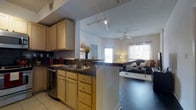 This single-floor residence offers an open-concept layout that connects the kitchen and living area in a cohesive, functional design. The kitchen is well-appointed with warm honey-toned cabinetry providing ample upper and lower storage, dark granite countertops, a stainless steel sink, and a stainless steel dishwasher. A peninsula countertop serves as both a prep space and a natural divider between the kitchen and living area, creating an inviting flow throughout. Track lighting in the kitchen ensures excellent task illumination, while tile flooring adds a clean, practical foundation underfoot. The living area transitions to rich, dark hardwood-style flooring, lending warmth and contrast to the open space. A ceiling fan centered in the living room provides both comfort and casual style. Natural light pours in through large windows dressed with light, sheer curtains, brightening the room and creating an airy atmosphere. The overall layout encourages easy interaction between the kitchen and living spaces, making it ideal for everyday living and entertaining alike. The neutral color palette throughout keeps the space feeling fresh, cohesive, and welcoming.