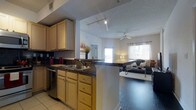 This single-floor residence offers an open-concept layout that connects the kitchen and living area in a cohesive, functional design. The kitchen is well-appointed with warm honey-toned cabinetry providing ample upper and lower storage, dark granite countertops, a stainless steel sink, and a stainless steel dishwasher. A peninsula countertop serves as both a prep space and a natural divider between the kitchen and living area, creating an inviting flow throughout. Track lighting in the kitchen ensures excellent task illumination, while tile flooring adds a clean, practical foundation underfoot. The living area transitions to rich, dark hardwood-style flooring, lending warmth and contrast to the open space. A ceiling fan centered in the living room provides both comfort and casual style. Natural light pours in through large windows dressed with light, sheer curtains, brightening the room and creating an airy atmosphere. The overall layout encourages easy interaction between the kitchen and living spaces, making it ideal for everyday living and entertaining alike. The neutral color palette throughout keeps the space feeling fresh, cohesive, and welcoming.