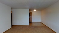 This single-floor space presents a straightforward and functional layout characterized by clean, neutral design elements. The main living area features wall-to-wall carpeting in a warm beige tone, providing a soft underfoot feel that extends throughout the space. The walls are finished in a light, neutral color, contributing to a bright and open atmosphere, while white baseboards frame the perimeter of the room cleanly.

The ceiling features a textured finish and is equipped with a flush-mount ceiling light fixture, providing centralized overhead illumination. The layout includes a partial wall that creates a subtle visual division within the open floor plan, adding a degree of structural interest to the otherwise open arrangement.

A notable feature is a freestanding wooden cabinet with decorative panel detailing, positioned near a doorway that leads to an adjacent area. The cabinet's warm, medium-toned wood finish adds a traditional element and serves as a functional storage piece within the space. Electrical outlets are positioned at standard intervals along the walls. Overall, the space conveys a clean, uncluttered environment well-suited for practical, everyday living.
