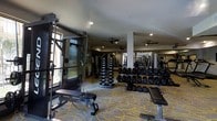 This fitness center is a thoughtfully designed workout facility that offers a wide range of equipment to support diverse training needs. The space features a generous free weights section, complete with multiple dumbbell racks holding an extensive selection of weights, as well as flat and adjustable benches for various exercises. A functional cable training machine anchors one side of the room, providing versatile resistance training options. The floor is covered in a durable, patterned carpet that adds a stylish touch while providing comfort underfoot. Ceiling fans are mounted throughout, ensuring consistent airflow and a comfortable environment during intense workouts. Recessed lighting illuminates the space evenly, creating a bright and energizing atmosphere. Full-length mirrors line the walls, allowing users to monitor their form and making the room feel more expansive. Structural columns are integrated neatly into the layout, preserving the open feel of the floor plan. The overall ambiance is modern and well-maintained, conveying a professional gym environment suited for both casual exercisers and dedicated fitness enthusiasts alike.