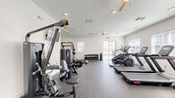 Located at 975 Pitt St in Clermont, FL, this dedicated fitness facility offers a thoughtfully organized exercise room across a single floor. The space is designed to accommodate a full range of workout activities, making efficient use of its floor area to provide a comfortable and functional environment.

The interior features crisp white walls and durable rubber-flooring throughout, creating a clean, professional gym atmosphere. Multiple ceiling fans and recessed lighting ensure the space remains well-ventilated and brightly lit. Several windows along the walls allow natural light to filter in, enhancing the overall environment.

The cardio section is equipped with multiple treadmills, an elliptical trainer, a stationary recumbent bike, and a rowing machine, providing diverse options for aerobic exercise. A wall-mounted television is positioned for viewing during cardio workouts. The strength training area features two multi-functional cable and weight machines, a Smith machine with an adjustable bench, and a well-stocked dumbbell rack ranging across multiple weight increments. A dedicated weight plate storage rack keeps the space organized. Together, these elements create a comprehensive, fully equipped fitness facility suitable for a wide variety of training goals.