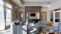 This single-floor space showcases a contemporary design with a carefully curated aesthetic that balances comfort and sophistication. The focal point of the room is a striking built-in media wall, featuring a wall-mounted flat-screen television framed by textured stone tile accents and warm wood paneling. Below the screen, dark cabinetry with a sleek countertop provides ample storage and display space, lending a refined, built-in bar-like quality to the area.

The seating arrangement is thoughtfully composed, with plush gray sectional sofas and accent chairs upholstered in complementary tones of silver and warm orange, creating an inviting and visually dynamic atmosphere. A round coffee table anchors the central seating area, adding an organic contrast to the room's clean architectural lines.

Recessed lighting and a tray ceiling detail contribute to the layered, polished look of the space, while natural light filters in through a doorway, brightening the neutral taupe and gray color palette. Framed wall art and fresh floral arrangements add finishing touches that enhance the space's welcoming, upscale ambiance.