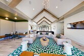 Located at 1580 Woodland Way in Okemos, Michigan, this generously sized single-story community facility delivers an impressive range of amenities across a sprawling floor plan. The centerpiece of the interior is a soaring vaulted great room defined by exposed wood beams, cathedral ceilings, and an elegant branching chandelier. Plush sectional seating is arranged atop a bold green-and-white geometric rug, while a stone fireplace and floor-to-ceiling windows flood the space with natural light and views of the surrounding greenery. A shuffleboard table anchors a separate lounge area, providing an inviting spot for recreation and socializing. Adjacent to the main lounge is a stylish coffee bar featuring a large island with bar seating, pendant lighting, and a playful wall graphic, all finished with light hardwood flooring and an arched accent wall. The fitness center is a standout feature, boasting vibrant orange ceilings, arched mirror alcoves, ceiling fans, and a comprehensive array of cardio and strength equipment, including a dedicated flex studio with large windows and natural light. Two bathrooms and a closet round out the interior. Outdoors, a resort-style pool with an attached spa, lounge chairs, and a covered patio with a metal roof creates a polished and welcoming recreational retreat.