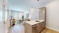 Located at 15410 S Brentwood St in Olathe, KS, this thoughtfully designed one-bedroom, two-bathroom unit offers a modern aesthetic with clean lines and high-quality finishes throughout. The open-concept living and kitchen area serves as the heart of the space, where light wood-tone flooring, recessed lighting, and large windows create a bright, welcoming atmosphere with outdoor views.

The kitchen is outfitted with stainless steel appliances, including a refrigerator, range, and microwave, complemented by warm, flat-front cabinetry and a generous island featuring a white quartz countertop, undermount sink, and bar seating — ideal for casual dining or entertaining. A sleek linear pendant light anchors the kitchen visually.

The living area offers comfortable proportions, with large windows allowing ample natural light and views of the surrounding area. The bedroom is carpeted for comfort, features a ceiling fan, and is served by two doors — one leading to a walk-in closet with built-in shelving and an in-unit washer/dryer, and another providing en-suite bathroom access. The primary bathroom includes a tub/shower combination with large-format white tile surround. A second full bathroom serves guests. The overall ambiance is clean, contemporary, and efficiently functional.