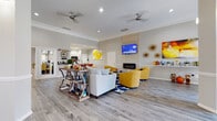 Located at 6147 Winged Elm Ct in Charlotte, NC, this single-floor community clubhouse is thoughtfully designed to serve as a welcoming social hub. The space is defined by soaring ceilings, crown molding, and light gray luxury vinyl plank flooring that flows seamlessly throughout.

The grand entrance foyer greets visitors with two sets of glass-paneled doors, large windows that flood the space with natural light, and a pair of vibrant yellow accent chairs — setting an inviting and energetic tone from the moment you walk in.

The main lounge area is a standout feature, anchored by a sleek linear fireplace set within a shiplap-clad accent wall, above which a wall-mounted television is positioned. Plush yellow swivel chairs arranged around a bold blue geometric area rug create a comfortable and stylish gathering space. Ceiling fans add to the comfort of the room, while colorful abstract artwork and decorative wall accents contribute to the lively aesthetic.

Adjacent to the lounge is a well-appointed coffee and snack bar, complete with a Keurig coffee station, counter seating with bold orange leather barstools, and a clean white countertop — ideal for casual conversation or a quick break.

A glass-partitioned corridor connects to a dedicated fitness room, which is fully visible through floor-to-ceiling glass panels, promoting transparency and an open feel throughout the entire facility.
