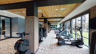 This single-story fitness center located on W Airport Blvd in Stafford, Texas, offers a thoughtfully designed workout environment that balances functionality with contemporary style. The space is organized into distinct zones, each tailored to a specific type of training. A long cardio corridor runs along one side, lined with treadmills, ellipticals, and rowing machines positioned in front of expansive floor-to-ceiling windows. These large glass panels frame outdoor views of a landscaped courtyard and allow abundant natural light to fill the space throughout the day. Exposed brick accent walls complement the warm wood-paneled ceilings, which feature recessed lighting and ceiling fans for comfort. Structural columns clad in wood-toned panels add architectural character while defining each section of the floor plan. The strength training area includes cable machines, a squat rack with bench, free weights, and battle ropes, providing a comprehensive setup for resistance-based workouts. A dedicated studio space houses spin bikes and a large display screen for guided classes, with full-length mirrors enhancing the sense of openness. Stylish storage cabinetry and custom signage add a polished, branded feel to the overall environment. The patterned carpet flooring ties the zones together cohesively.