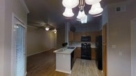 This single-floor residence offers an open and functional layout that seamlessly connects the kitchen and living areas. The kitchen is a standout feature, equipped with warm-toned wood cabinetry that provides ample storage, a full suite of black appliances including a range, microwave, and refrigerator, and a central island with a built-in sink and light countertop surface — ideal for meal preparation and casual dining. An elegant multi-light chandelier hangs above the island, adding a decorative focal point to the space. The kitchen flows naturally into the adjoining living area, which is finished with hardwood-style flooring and features a ceiling fan with integrated lighting, contributing to a comfortable and well-ventilated atmosphere. Crown molding details along the upper walls lend a polished, refined character to the interior. The entryway, visible from the main living space, provides a clear sense of the home's open circulation and layout. Neutral wall tones and consistent flooring throughout create a cohesive aesthetic that is both inviting and easy to personalize. Overall, the space balances practicality with a warm, livable ambiance well-suited for everyday living.