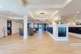 Located at Presidential Way in West Palm Beach, Florida, this generously sized single-floor common area spans an expansive footprint designed to serve as a welcoming community hub. The space is organized into distinct functional zones that flow seamlessly into one another, united by consistent light-toned hardwood flooring accented with dark inlay border detailing throughout.

The main lobby entry features glass double doors that frame views of the surrounding palm-lined exterior. Inside, tray ceilings with recessed lighting define different areas of the space, while a striking multi-ring circular chandelier serves as a focal point over the central corridor. Crisp white walls and classic white columns reinforce a clean, modern coastal aesthetic.

A prominent mailbox wall lined with dark-finished individual resident mail units runs along one side, topped with a display ledge adorned with potted plants and decorative accents. Multiple seating vignettes are arranged throughout the lobby, featuring high-back woven chairs and occasional tables that invite comfortable gathering.

Toward the interior, a dedicated lounge and bar area offers an extended white countertop with barstool seating, complemented by globe pendant lights and large glass doors opening to natural light beyond. The overall ambiance is bright, polished, and welcoming, with thoughtful details that balance functionality and style.