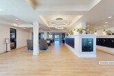 Located at Presidential Way in West Palm Beach, Florida, this generously sized single-floor common area spans an expansive footprint designed to serve as a welcoming community hub. The space is organized into distinct functional zones that flow seamlessly into one another, united by consistent light-toned hardwood flooring accented with dark inlay border detailing throughout.

The main lobby entry features glass double doors that frame views of the surrounding palm-lined exterior. Inside, tray ceilings with recessed lighting define different areas of the space, while a striking multi-ring circular chandelier serves as a focal point over the central corridor. Crisp white walls and classic white columns reinforce a clean, modern coastal aesthetic.

A prominent mailbox wall lined with dark-finished individual resident mail units runs along one side, topped with a display ledge adorned with potted plants and decorative accents. Multiple seating vignettes are arranged throughout the lobby, featuring high-back woven chairs and occasional tables that invite comfortable gathering.

Toward the interior, a dedicated lounge and bar area offers an extended white countertop with barstool seating, complemented by globe pendant lights and large glass doors opening to natural light beyond. The overall ambiance is bright, polished, and welcoming, with thoughtful details that balance functionality and style.