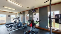 This single-floor fitness center offers a comprehensive workout environment designed to support a wide range of exercise routines. The space is divided into distinct workout zones, all finished with durable dark rubber flooring that provides traction and cushioning throughout. Warm wood crown molding trims the walls and windows, adding a polished architectural detail to the functional interior.

The cardio zone is well-stocked with multiple treadmills, an elliptical trainer, and a rowing machine, positioned alongside large windows that invite abundant natural light and offer views of the surrounding outdoor landscape. A wall-mounted flat-screen television is positioned for easy viewing during cardio sessions.

The strength and functional training area includes a Bowflex cable and pulley multi-gym system, a plyo box, a BOSU balance trainer, and a stability ball, offering versatile options for resistance and bodyweight training. Wall-mounted mirrors provide visual feedback during workouts.

The space is brightly lit with overhead fluorescent fixtures and benefits from multiple windows on several walls, ensuring a well-ventilated and open atmosphere. A dedicated entry door with access to the exterior, along with a water cooler station, adds everyday convenience. The overall layout is spacious and thoughtfully organized, making it suitable for simultaneous use by multiple individuals.