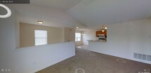 This single-floor home offers an inviting open-concept design that seamlessly connects the main living areas. The living space is distinguished by soaring vaulted ceilings that enhance the sense of openness and volume, while neutral-toned walls and light carpeting create a warm, welcoming atmosphere throughout. A half-wall partition adds subtle definition between the entry and living areas without disrupting the flow of the space. Natural light filters in through a window in the dining area, which is accented by an elegant chandelier that adds a decorative focal point overhead. The kitchen is positioned adjacent to the dining area and features warm honey-toned wood cabinetry, a light-colored countertop with a peninsula-style breakfast bar, a range with overhead ventilation hood, and tile flooring that transitions naturally from the carpeted living spaces. The open sightlines between the kitchen, dining, and living areas make this home well-suited for both everyday living and entertaining. The consistent use of soft, neutral finishes throughout lends the interior a cohesive and airy aesthetic, while the vaulted ceiling design adds architectural character and distinction to this single-level residence.