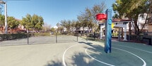 This outdoor recreational space centers on a full basketball court surfaced in green and gray with clearly marked regulation lines, including the key, three-point arc, and mid-court boundary. Two standard basketball hoops with backboards are positioned at each end of the court, providing a complete playing experience. The court is fully enclosed by a chain-link fence with gated entry points, ensuring a defined and secure play area.

A distinctive blue perforated equipment tower is positioned courtside, adding a functional and visual accent to the space. Mature deciduous and ornamental trees line the perimeter, offering partial shade and a natural buffer between the court and the surrounding residential buildings. The neighboring architecture features Mediterranean-influenced design elements, including tiled rooflines, visible in the background.

The court benefits from full exposure to natural light, making it well-suited for daytime use. The open-air setting, combined with the lush tree canopy and clear sightlines to the sky, creates an energetic yet pleasant atmosphere. The space is designed for active recreational use and serves as a communal amenity within its residential setting.