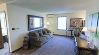 This single-floor space offers a warm and inviting atmosphere, centered around a well-appointed living area. The room features plush wall-to-wall carpeting that adds a sense of comfort underfoot, complemented by neutral-toned walls that create a bright and welcoming environment. Two windows allow natural light to fill the space, enhancing its open feel. A wall-mounted mini-split air conditioning unit ensures year-round climate control. The living area is designed for relaxation and functionality, with ample room for seating and décor. A large decorative mirror with a dark wood frame adorns one wall, adding depth and visual interest to the room. Warm accent lighting from a floor lamp contributes to the cozy ambiance. A bistro-style high table and chairs provide a casual seating option, while decorative stacked storage boxes add a charming, personalized touch. The space strikes a balance between practicality and comfort, making it a welcoming retreat suitable for everyday living.