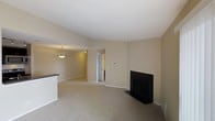 This single-floor space offers an open and airy layout with a warm, neutral color palette of soft beige tones on the walls and plush wall-to-wall carpeting underfoot. The living area is anchored by a classic black fireplace with a sleek surround, serving as a natural focal point in the room. The vaulted or angled ceiling adds a sense of height and spaciousness to the interior.

A partial view of a counter or bar-height surface is visible at the edge of the living space, suggesting a transition to a kitchen or dining area. A pendant light fixture hangs overhead in this adjacent zone, providing warm, focused illumination and adding a decorative touch. The open doorway visible in the background leads further into the unit, hinting at additional rooms beyond.

The overall ambiance is calm and understated, with clean lines and minimal architectural detail creating a versatile canvas. Natural and artificial lighting work together to brighten the space, while the neutral finishes throughout ensure a cohesive and inviting atmosphere well-suited for comfortable everyday living.