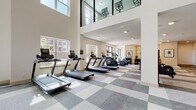 This fitness center is designed to support a full range of workout routines within a bright, open, and modern environment. The space is defined by its impressive double-height ceilings, which create an airy, expansive atmosphere throughout. Large windows along the perimeter flood the floor with natural light, offering views of the surrounding buildings while enhancing the sense of openness.

The cardio zone is well-stocked with multiple treadmills, elliptical machines, stair climbers, and stationary bikes — including both upright and recumbent models — arranged in organized rows across the patterned tile and carpet flooring. A mezzanine-level glass railing above adds architectural interest and visual depth to the main workout area.

The strength training section features a full rack of dumbbells, adjustable weight benches, a multi-functional cable machine by Precor, and a Beaver Fit storage unit, offering versatility for a wide variety of resistance exercises. A power rack is also available for barbell training.

Recessed lighting supplements the natural light throughout, ensuring the space remains well-lit at all times. The neutral color palette of white walls and soft grey flooring contributes to a clean, professional aesthetic. A dedicated stretch and cool-down area with seating rounds out the facility, making this fitness center a comprehensive and inviting space for all levels of fitness.